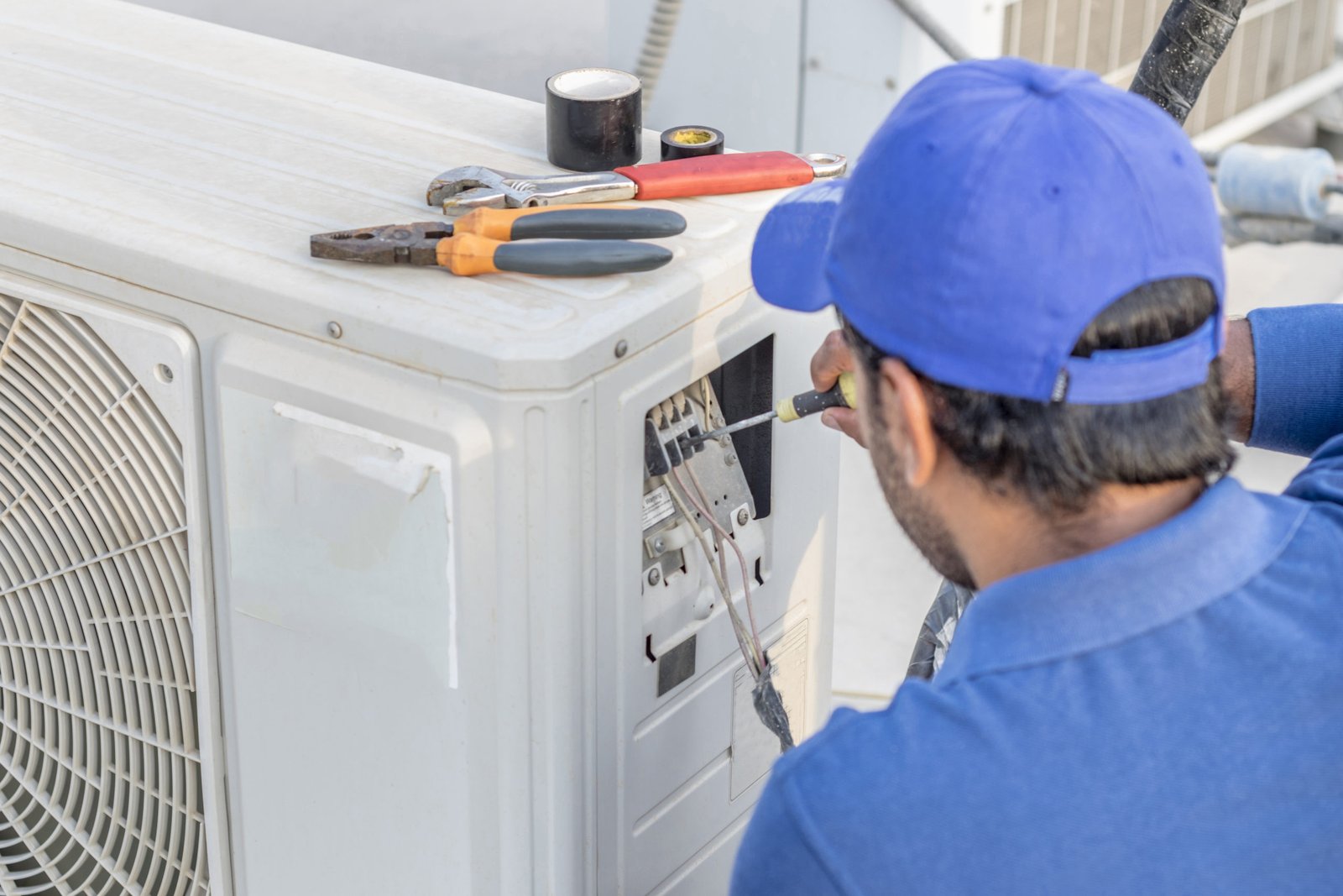 a professional electrician man is fixing a heavy duty unit of central air conditioning system by his tools on the roof top and wearing  uniform and white cap